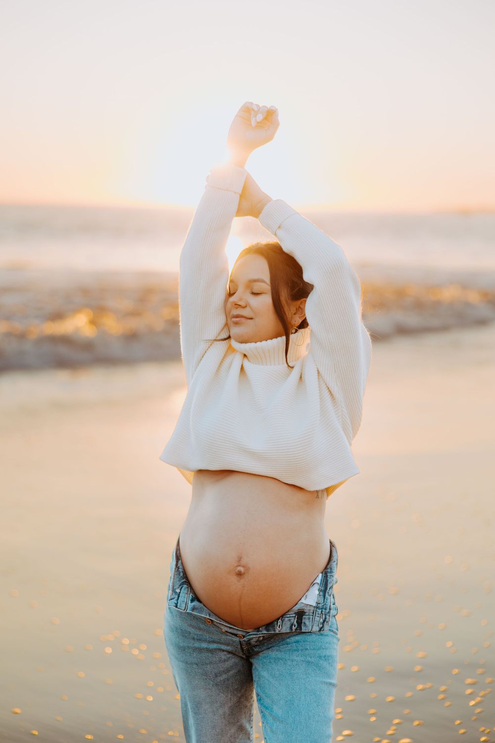 A pregnant woman in a white sweater and jeans stretches joyfully at sunset on a beach.