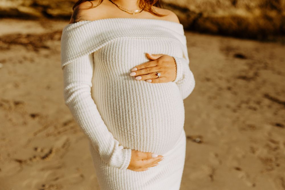 A pregnant woman in a white off-shoulder sweater dress cradles her baby bump while standing on sand.