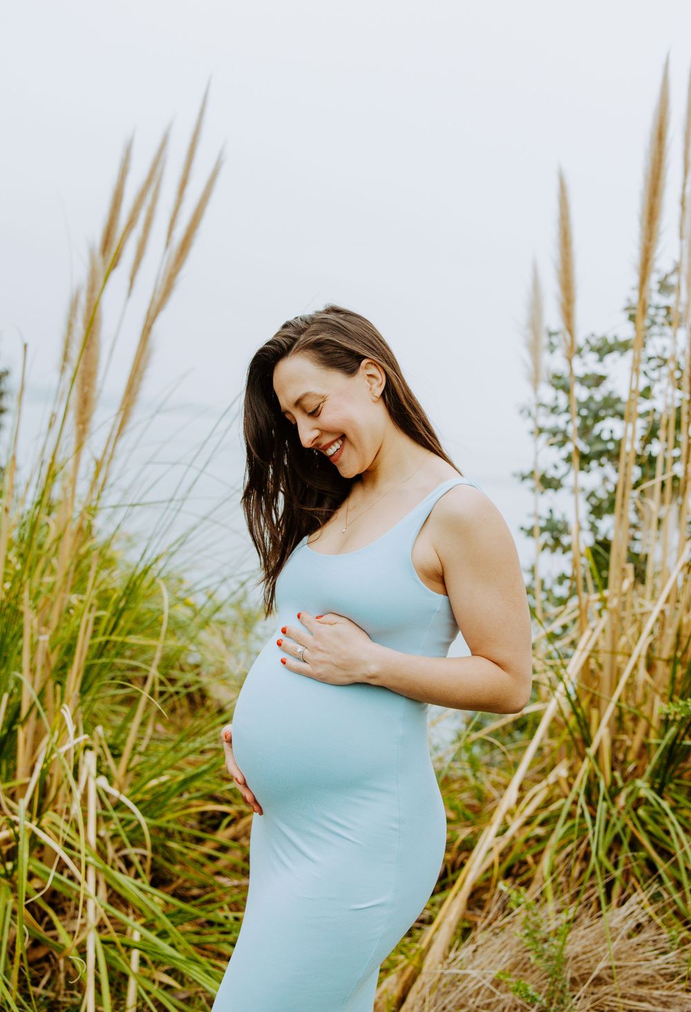 A radiant expectant mother in a light blue dress smiles while standing among tall beach grass on a sunny day.