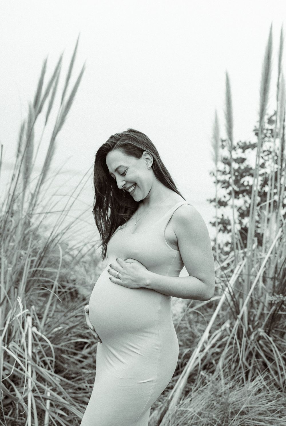 A black and white maternity photo shows a woman in a fitted dress surrounded by tall beach grass.
