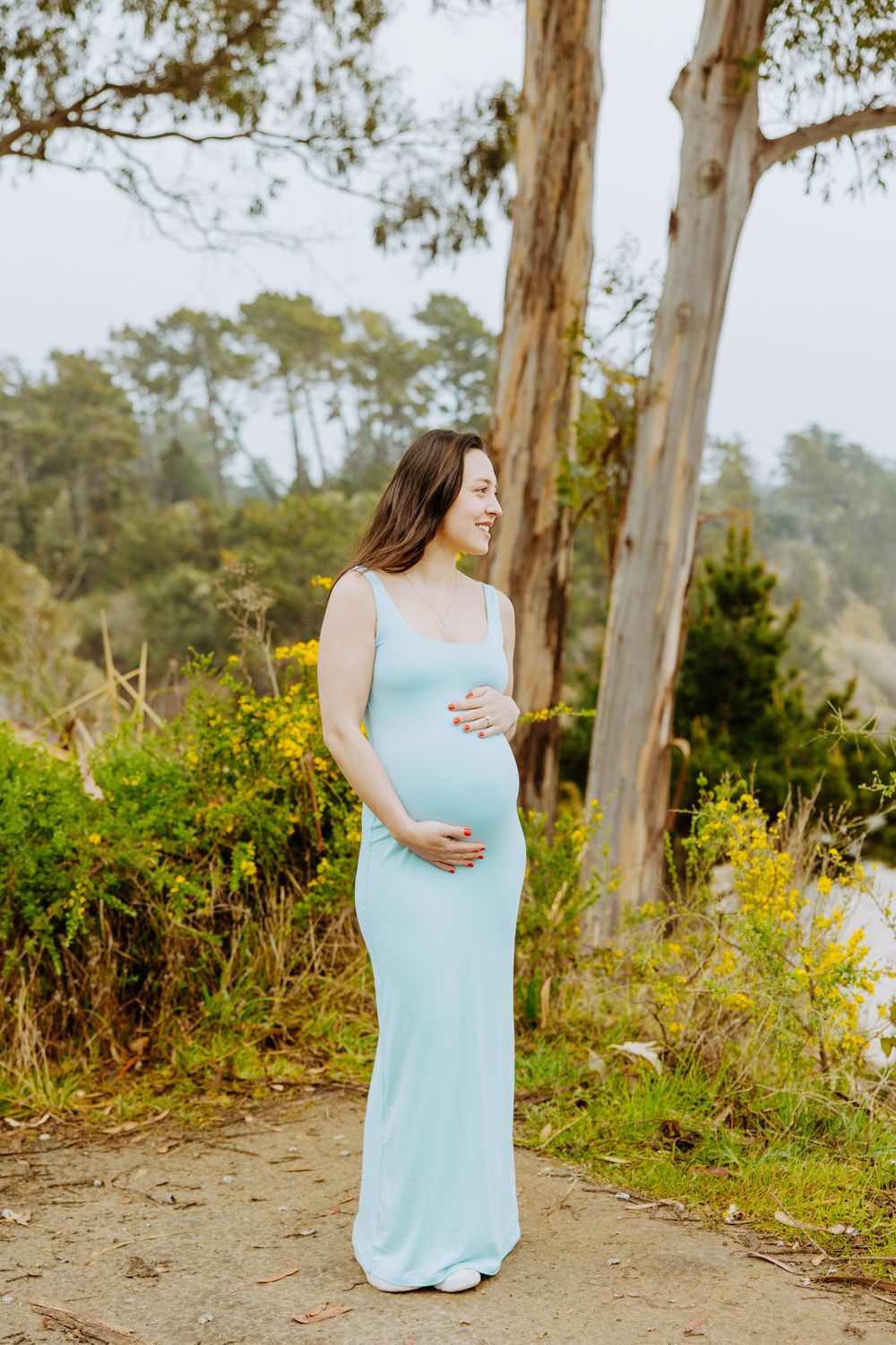 Expectant woman in light blue flowing maxi dress stands on nature trail surrounded by eucalyptus trees and greenery.