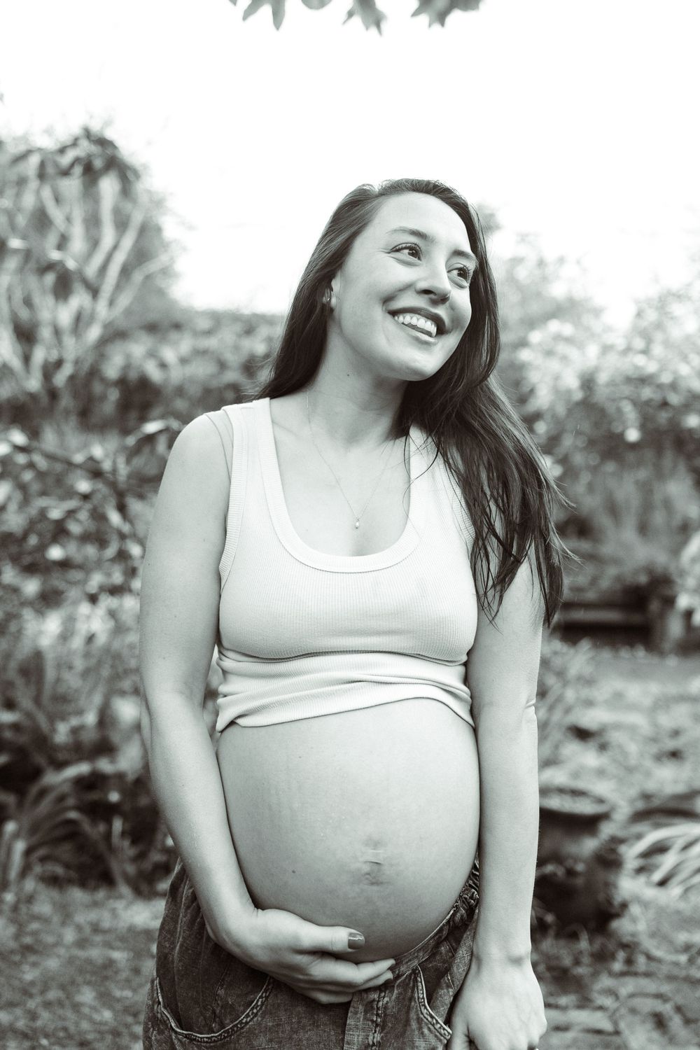 Black and white portrait of an expecting mother outdoors smiling and cradling her baby bump in a white tank top.