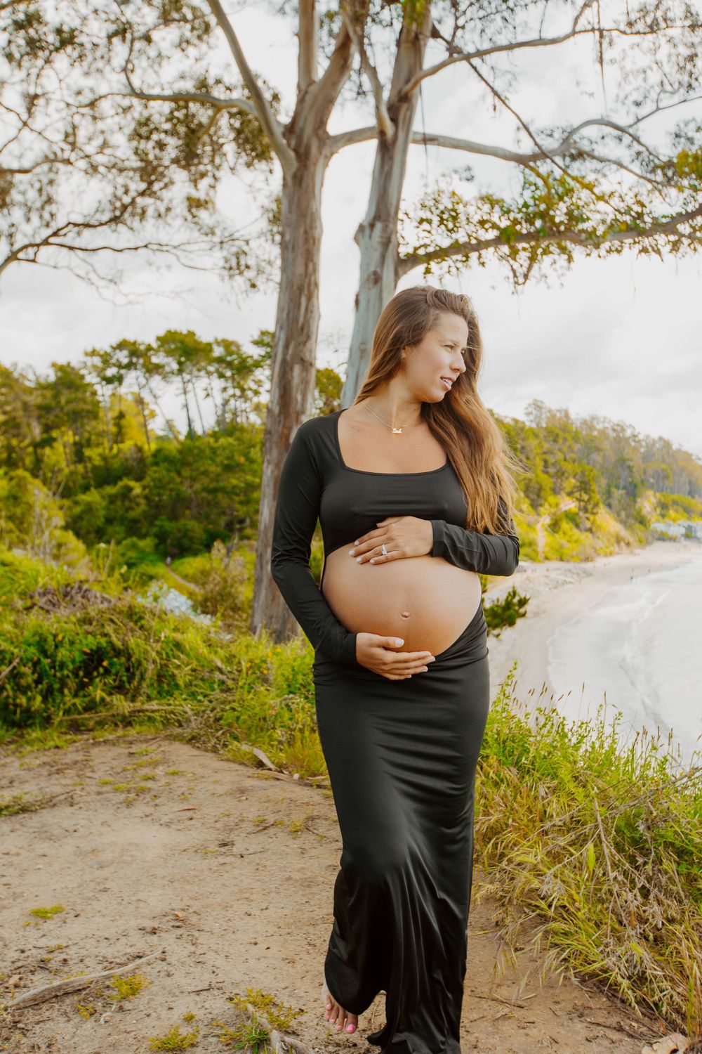 A pregnant figure in a long black dress stands on a dirt path near trees during golden hour.