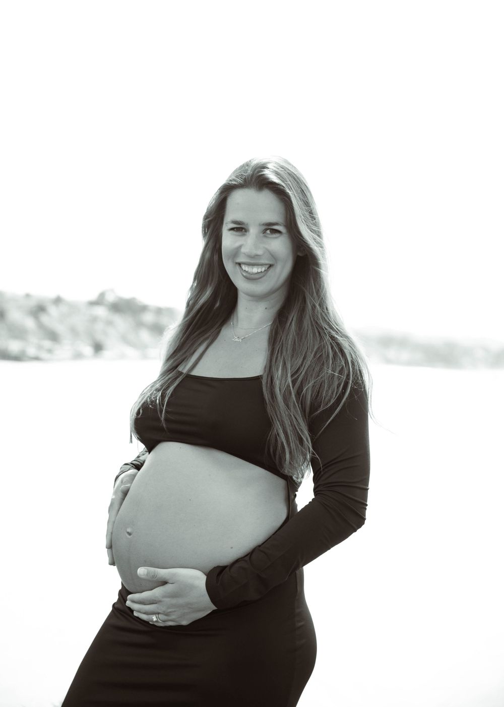 Black and white portrait of an expectant mother wearing a dark dress and cradling her baby bump outdoors.