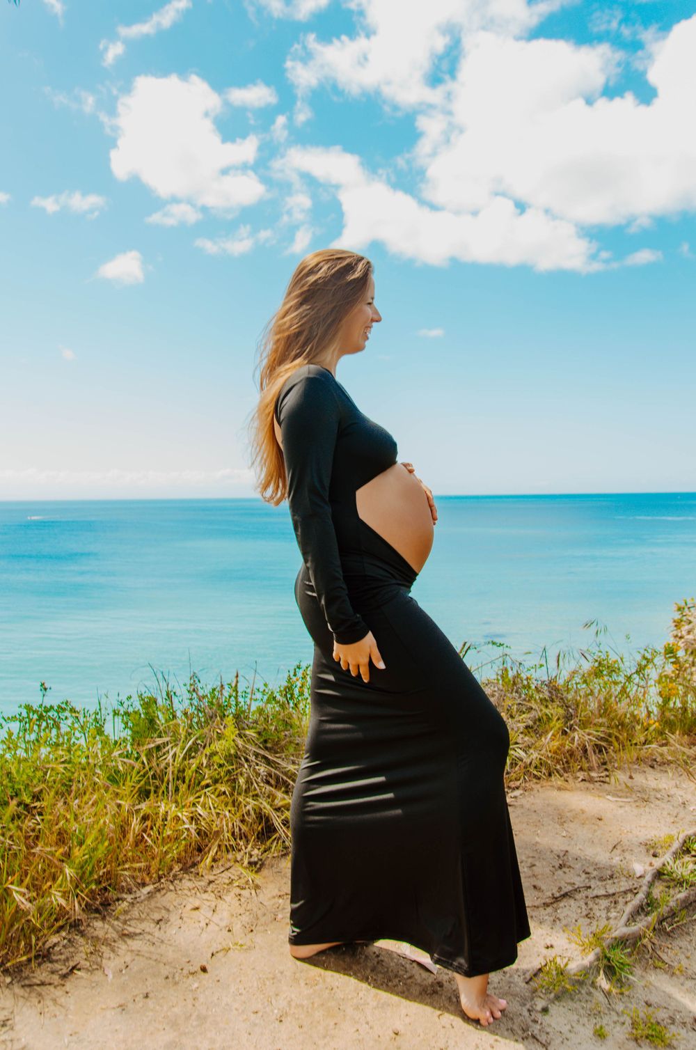 Pregnant woman in black dress stands on coastal cliff overlooking turquoise ocean waters under bright blue sky with clouds.