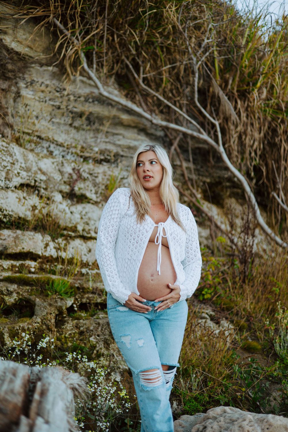 A pregnant woman in a white sweater and ripped jeans poses against a rocky cliff backdrop in nature.