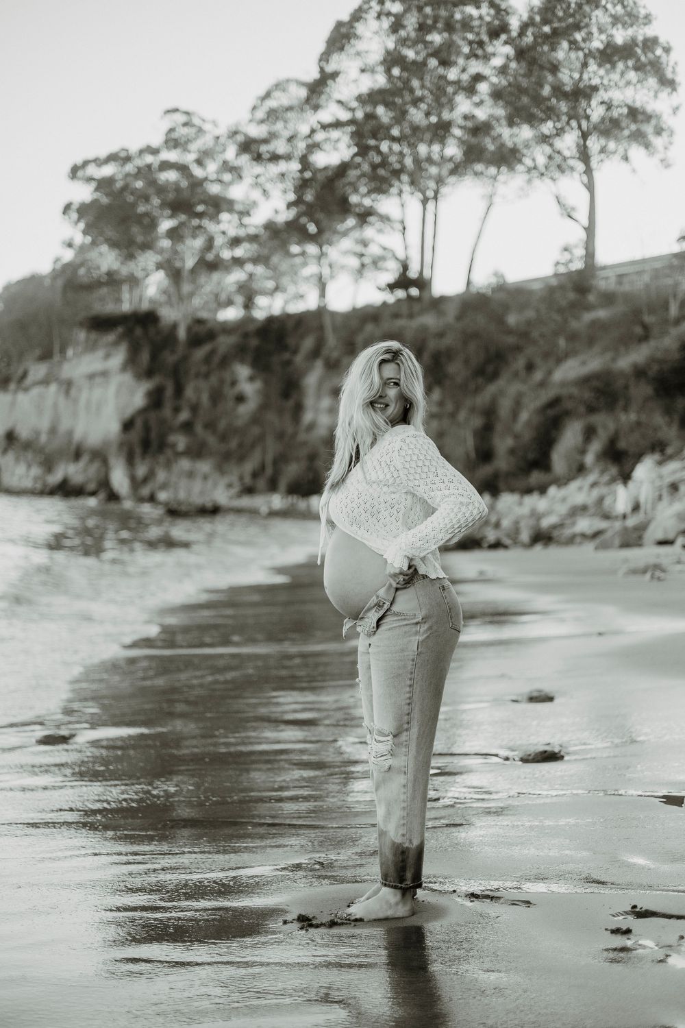 Black and white photo of person in knit sweater standing on misty beach with cliffs and trees in background.