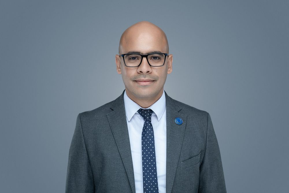 Professional headshot of person wearing black-rimmed glasses, charcoal suit and polka dot tie against gray background.