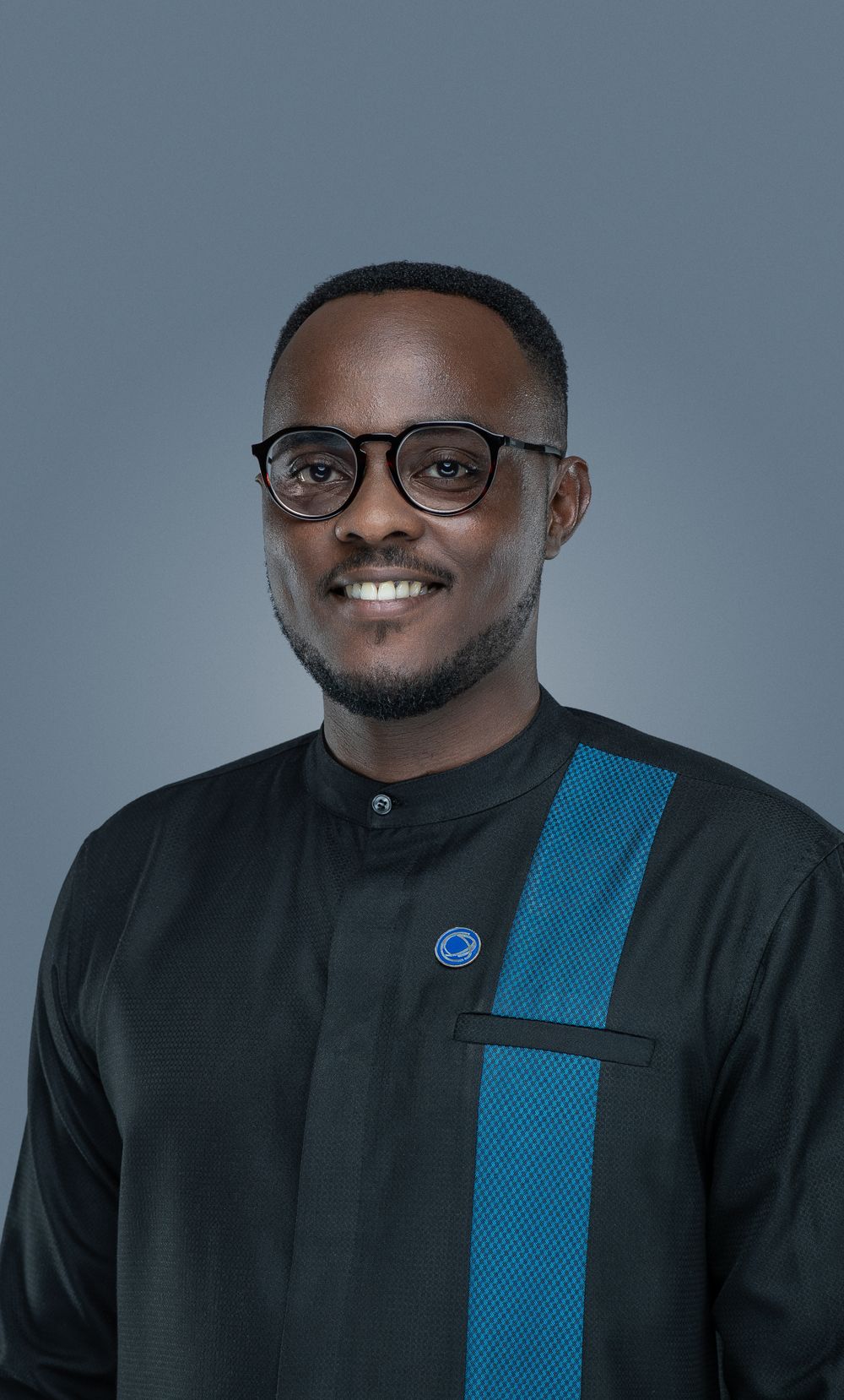 Smiling person wearing a black traditional-style shirt with blue trim detail and round glasses against a gray backdrop.