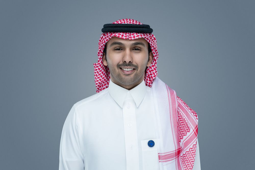 A person wearing traditional Middle Eastern dress including a white thobe and red and white checkered keffiyeh against a gray background.