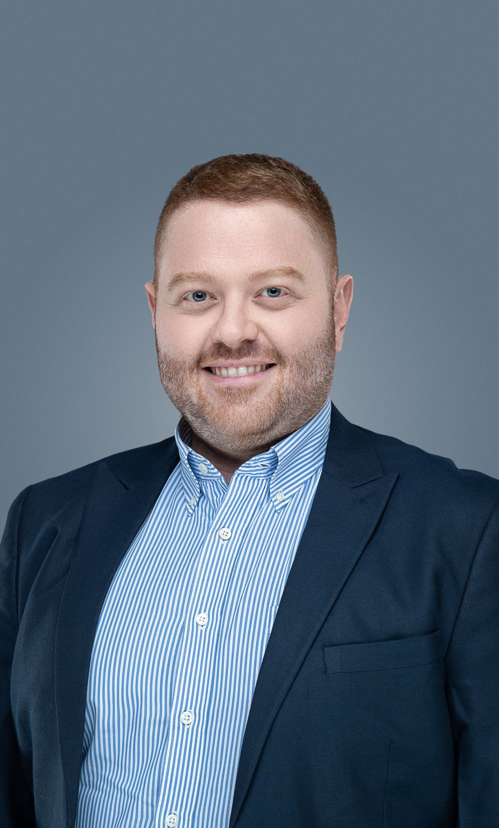 Professional headshot of a smiling person with a beard wearing a navy blazer and striped button-down shirt against a gray backdrop.