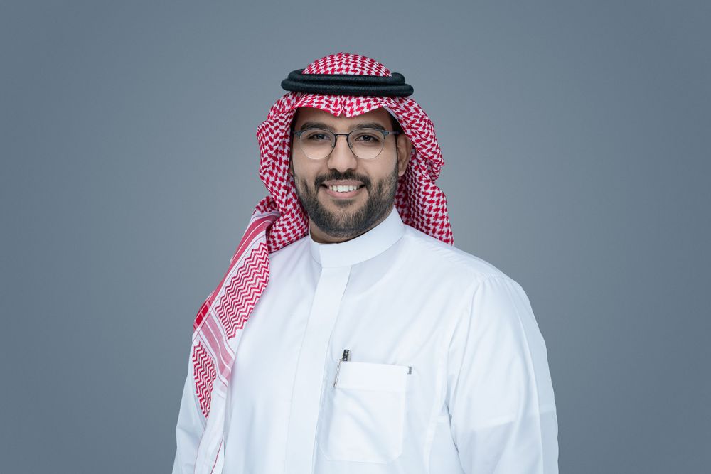 A person wearing traditional Middle Eastern thobe and red checkered keffiyeh smiles against a gray background.