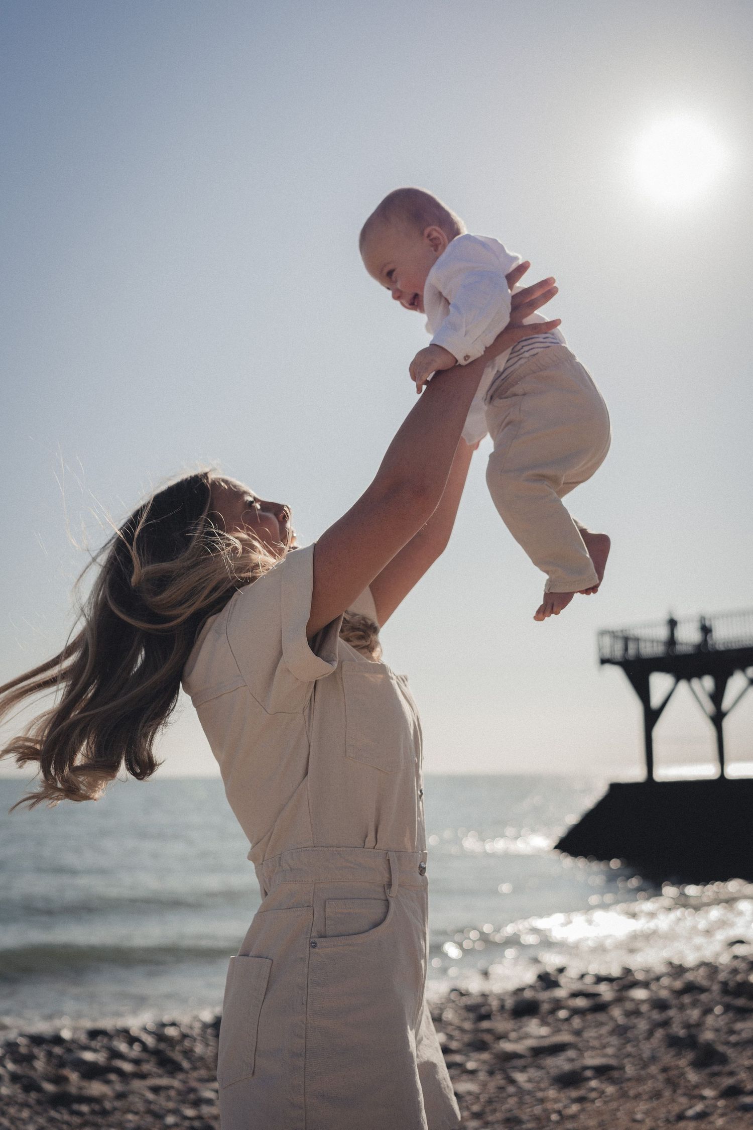 Parent soulevant son bébé devant un paysage ensoleillé en bord de mer, avec une jetée en arrière-plan.