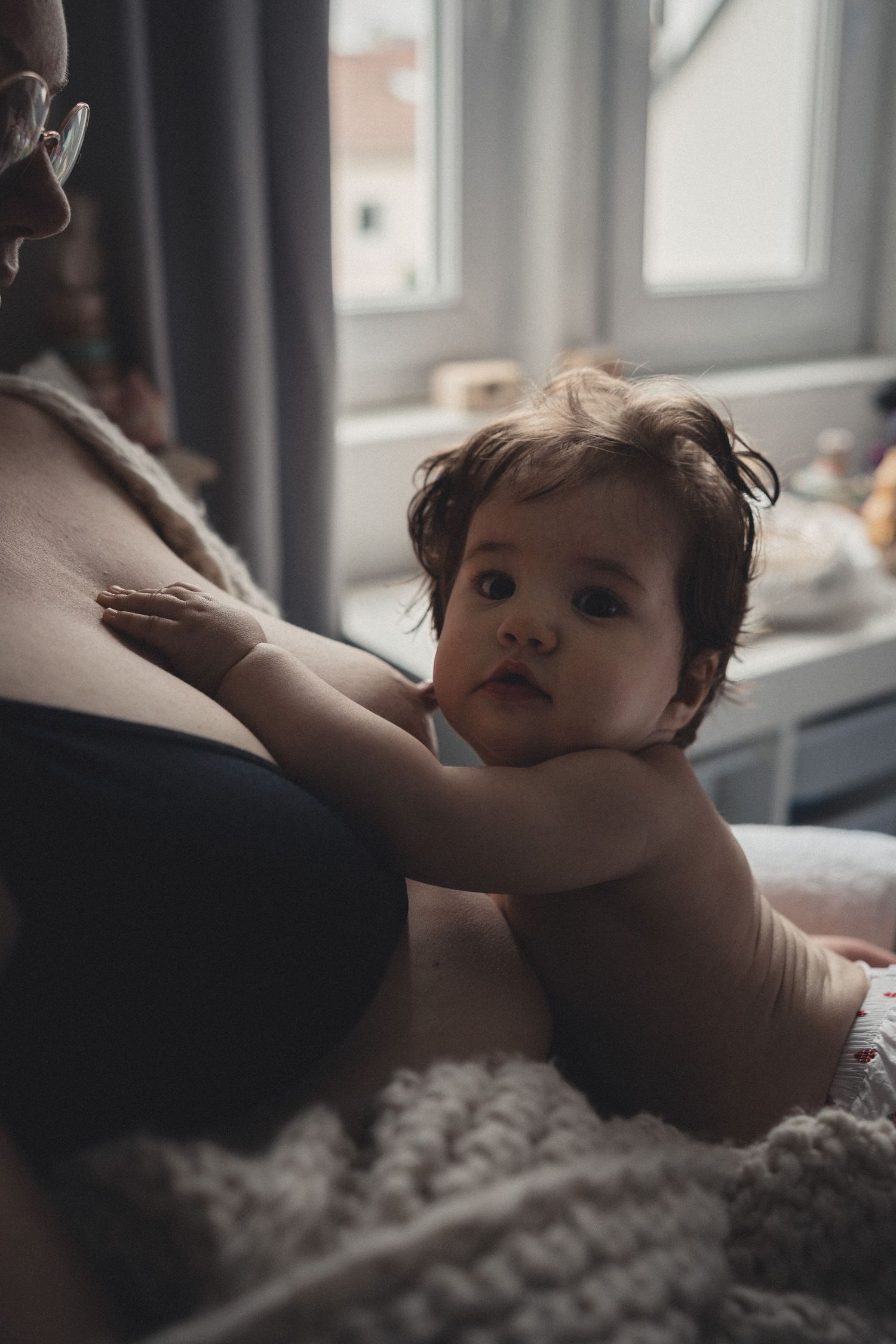 Baby in diaper reaching up while resting on soft bedding near a window.
