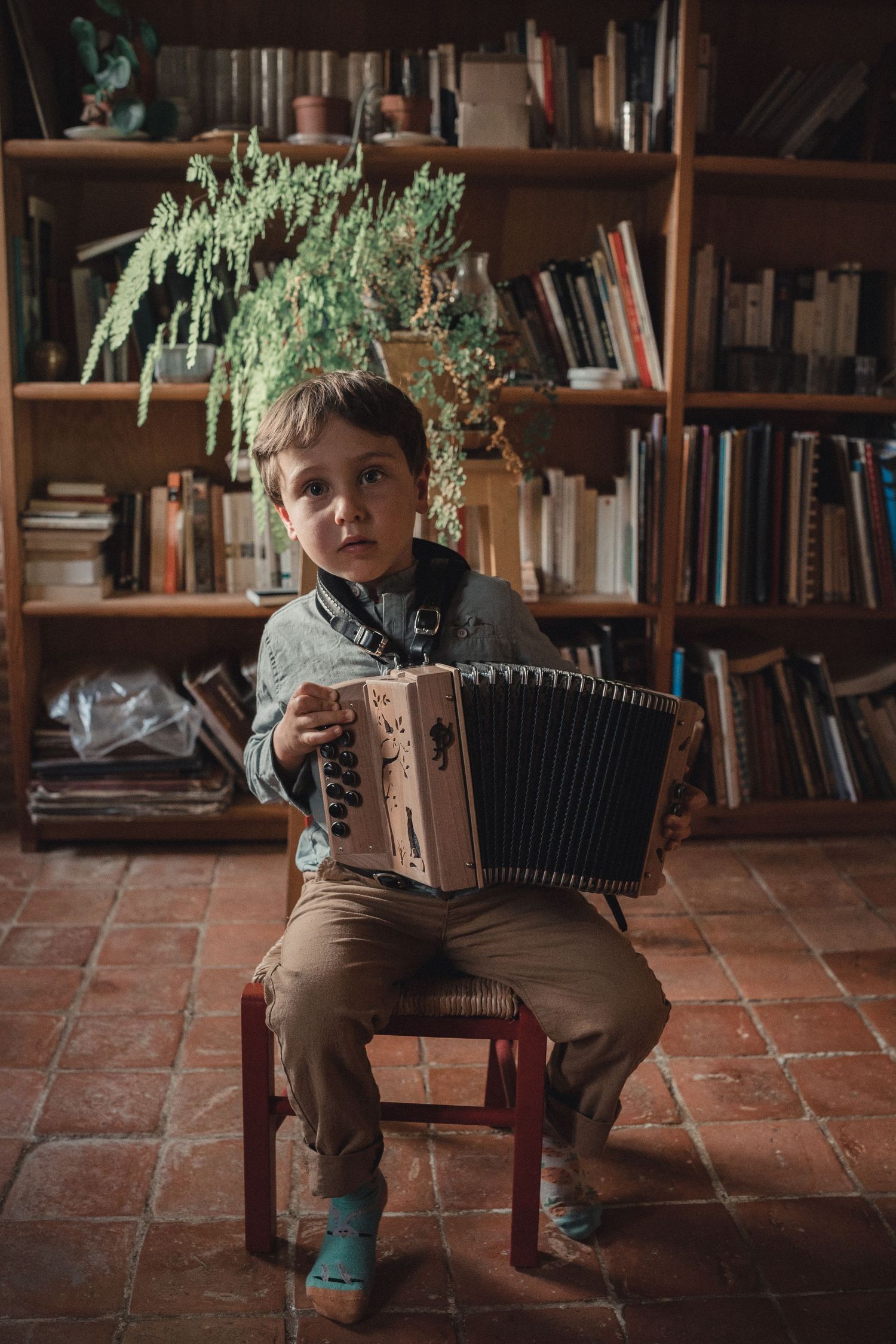 Jeune enfant assis sur une chaise en bois, jouant de l’accordéon devant une étagère remplie de livres.