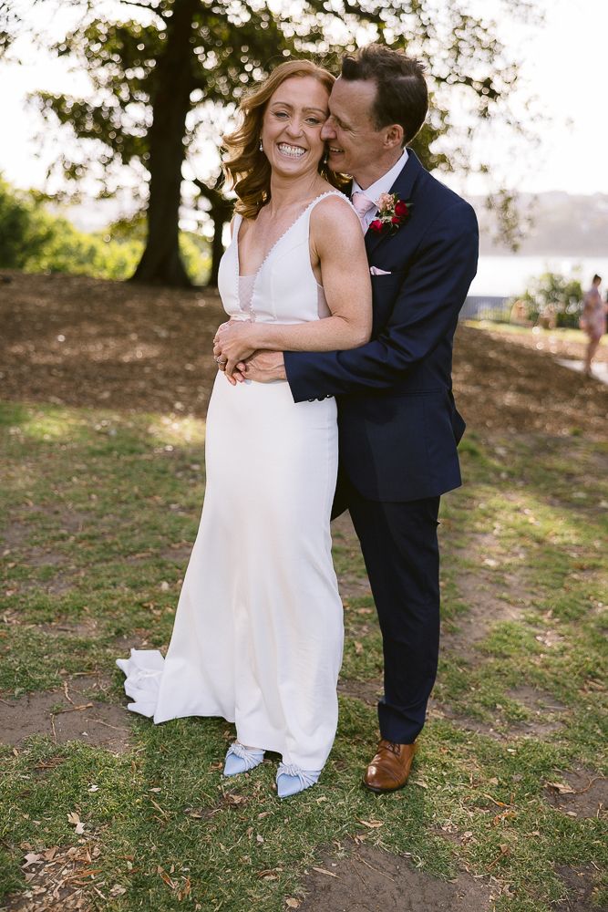 A couple embraces in a romantic outdoor wedding setting with trees and water in the background.