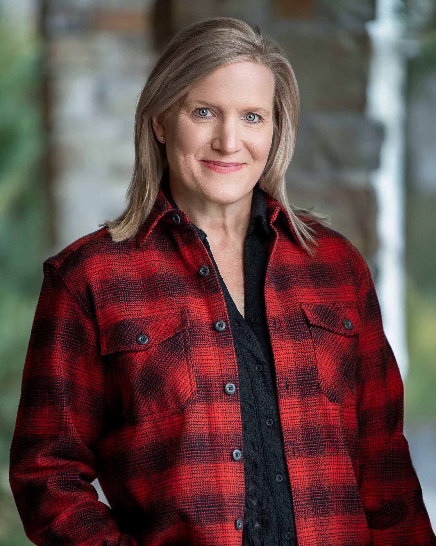 Woman wearing red and black buffalo plaid jacket poses in soft natural lighting with blurred forest background.