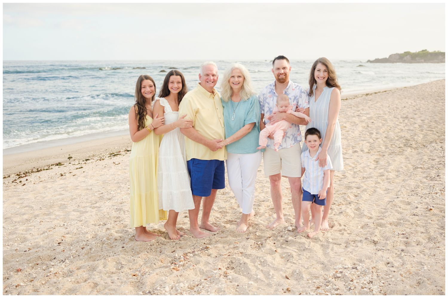 Group in coordinating blue and yellow outfits posing together on a sunny beach with ocean waves in background.