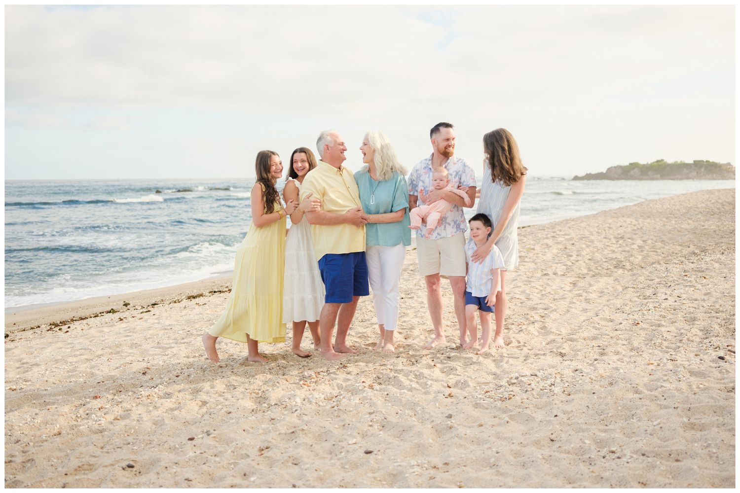 A family group wearing pastel colors stands together and smiles on a sunny beach by the ocean waves.