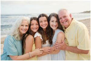 A happy family group embraces while standing together on a sunny beach with ocean waves in the background.