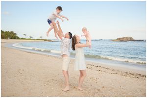 Family enjoying a playful moment on a sandy beach with ocean waves and rocky formations in background.