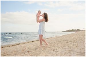 A mother in a light blue dress lifts baby into the air on a sunny beach with waves crashing in the background.