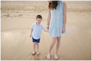 A child and adult wearing light blue outfits hold hands while standing barefoot on a sandy beach.