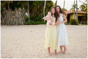 Two friends in summery dresses stand smiling together on a tropical beach with palm trees and beachwalls in the background.
