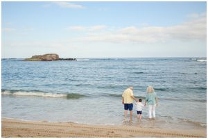 Three people stand at the edge of the ocean waves on a sandy beach with a rocky island in the distance.