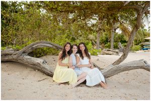 Three friends in light summer dresses sit together on a fallen tree at a tropical beach with palm trees in the background.
