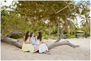 Three friends sit together on a fallen tree branch at a tropical beach with palm trees and soft sand.