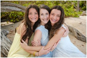 Three people wearing pastel dresses embrace while sitting on rocks at the beach on a sunny day.
