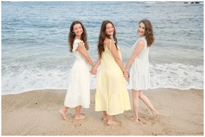 Three women in flowy summer dresses pose together on a beach shore with ocean waves in the background.