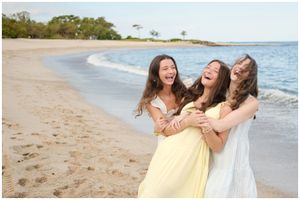 Three friends embrace and laugh together on a sandy beach with the ocean and trees in the background.