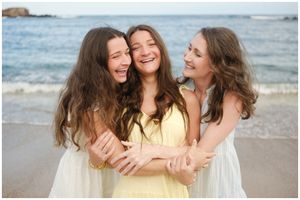 Three friends share a joyful embrace while laughing together on a sunny beach with ocean waves in the background.