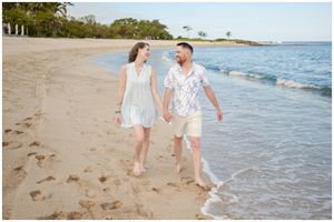 Two people walking hand in hand along a tropical beach shoreline in light summer attire.