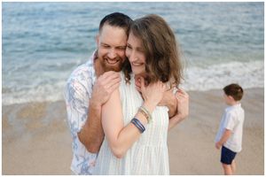 A couple share a joyful moment on the beach while a child stands in the background near the ocean.