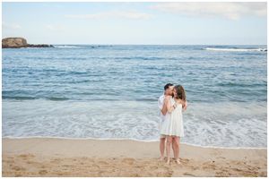 Two figures in white dresses embrace on a sandy beach with ocean waves and rocks in the background.