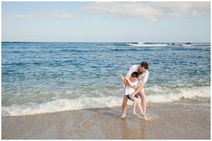 A romantic couple embraces at the edge of ocean waves on a sunny beach with blue water in the background.