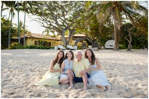 A group relaxes together on a sandy beach with palm trees and houses in the background on a sunny day.