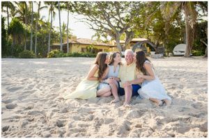 Three people sitting together playfully on a sandy tropical beach with palm trees and buildings in the background.