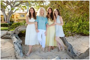 A group of people in pastel colored dresses stand together on driftwood at a beach in warm sunlight.