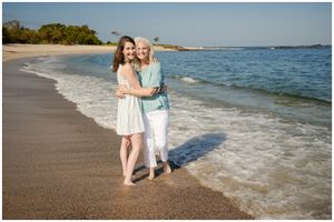 Two people embrace while standing on a sandy beach with turquoise ocean waters and coastline in the background.