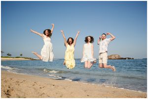 Four people in white and yellow dresses jumping joyfully on a scenic beach with blue waters and clear sky.