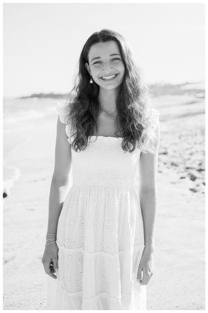A woman in a white eyelet dress stands on a beach in black and white photography.