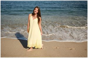 A person in a flowing yellow sundress stands barefoot on a sandy beach with ocean waves in the background.