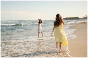 Two people in flowing dresses walking along the shoreline at sunset as waves roll onto the sandy beach.