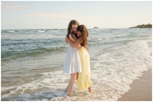 Two people in summer dresses embrace while standing in ocean waves on a sunny beach.