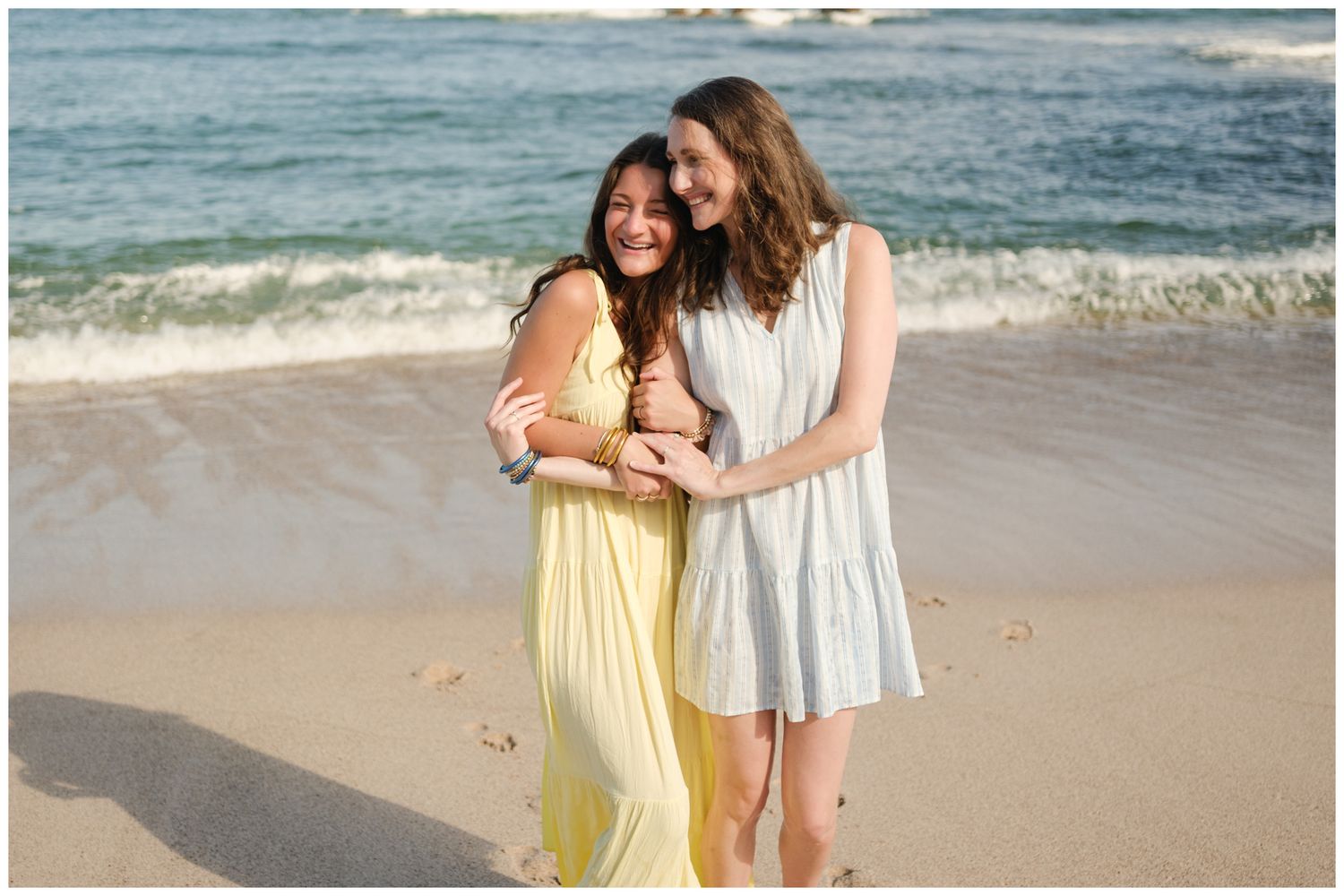 Two friends embrace while standing on a sandy beach with ocean waves in the background.