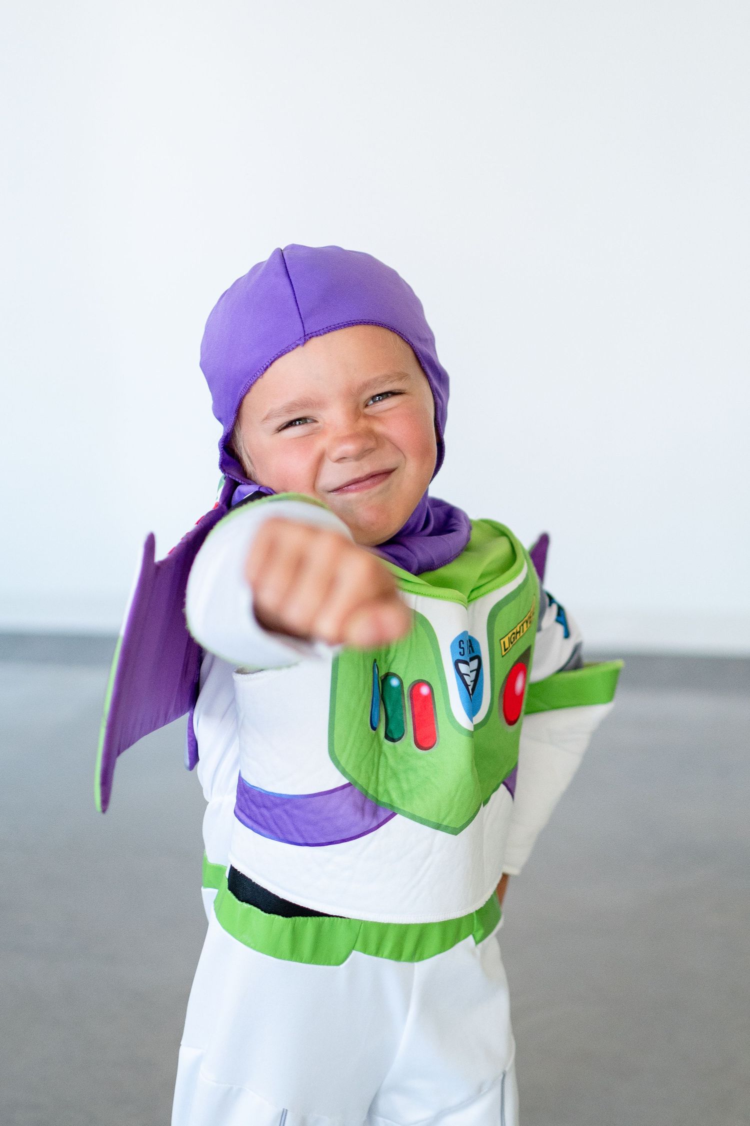 Small child dressed in a Buzz Lightyear costume with white and green suit and purple cap strikes a playful pose.